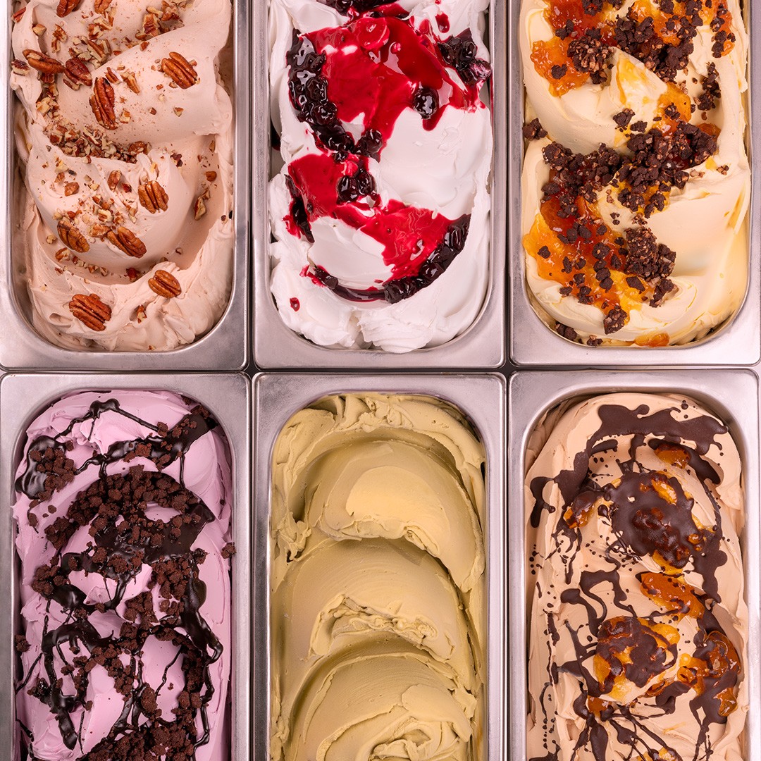 Seis bandejas metálicas de gelato em sabores variados, incluindo pecã, frutas vermelhas, pistache e chocolate com car...